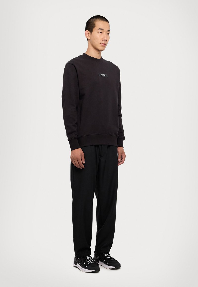 Felpa nera a girocollo con un'etichetta rettangolare sul petto, abbinata a pantaloni neri larghi e sneaker nere. Texture liscia, design casual.