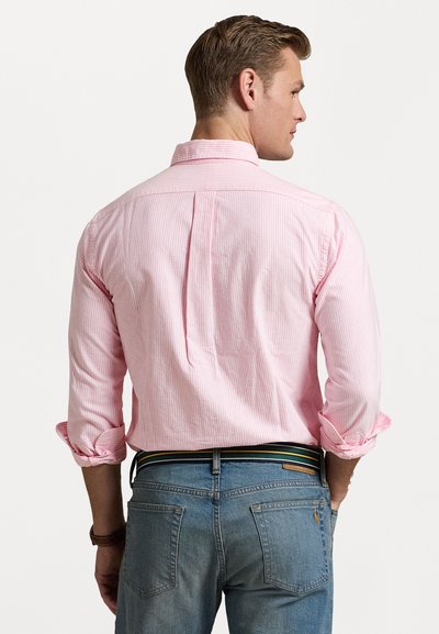 Camisa riscada em rosa claro com colarinho abotoado, mangas longas arregaçadas e pregas nas costas, combinada com jeans denim azuis e um cinto multicolorido.