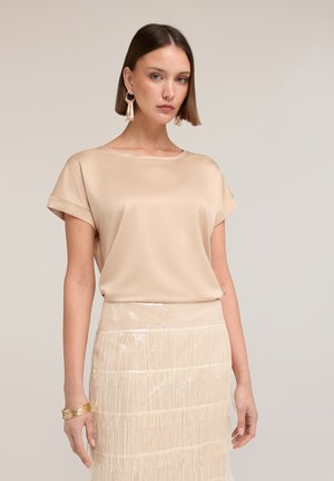 Vrouw met steil bruin haar draagt een beige blouse met korte mouwen, een gelaagde franjes rok, gouden creolen en meerdere gouden armbanden aan haar linkerpols.