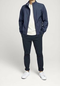 Marineblauwe zip-up jack met een hoge kraag, grijs sweatshirt eronder, gecombineerd met marineblauwe broeken en witte sneakers. Geweven stof, minimalistisch ontwerp.