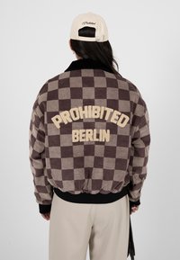 Personne portant une veste à carreaux marron et beige avec l'inscription « PROHIBITED BERLIN » dans le dos, une casquette beige et un pantalon clair, vue de dos.