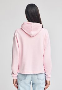 Rosa munkjacka med huva, långa ärmar, mjuk textur och kort design, tillsammans med ljusblå jeans. Synliga sömmar och huvdetaill.