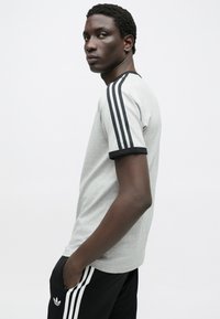 Γκρι κοντομάνικη μπλούζα με μαύρες λεπτομέρειες στις μανσέτες και τρεις μαύρες ρίγες σε κάθε ώμο, συνδυασμένη με μαύρο παντε�λόνι με λογότυπο.