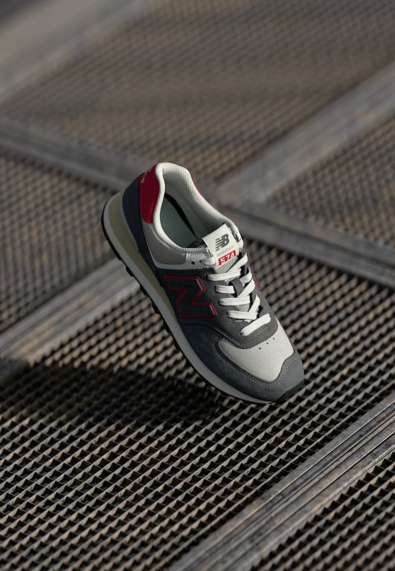 Grå og rød New Balance sneakers med en overdækning af stof og ruskind, tekstureret sål, hvide snørebånd og et stort logo på siden.