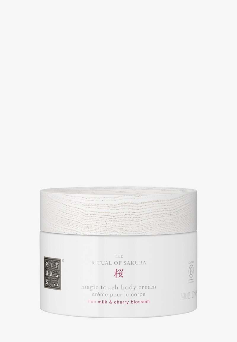 Biały cylindryczny pojemnik z teksturowaną pokrywką przypominającą drewno. Etykieta zawiera różowy napis "Magic Touch Body Cream, Mleko ryżowe i Kwiat Wiśni."