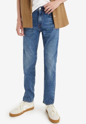 Personne portant un jean bleu droit, des baskets blanches avec semelles en gomme, une chemise blanche et une chemise marron ouverte à boutons.