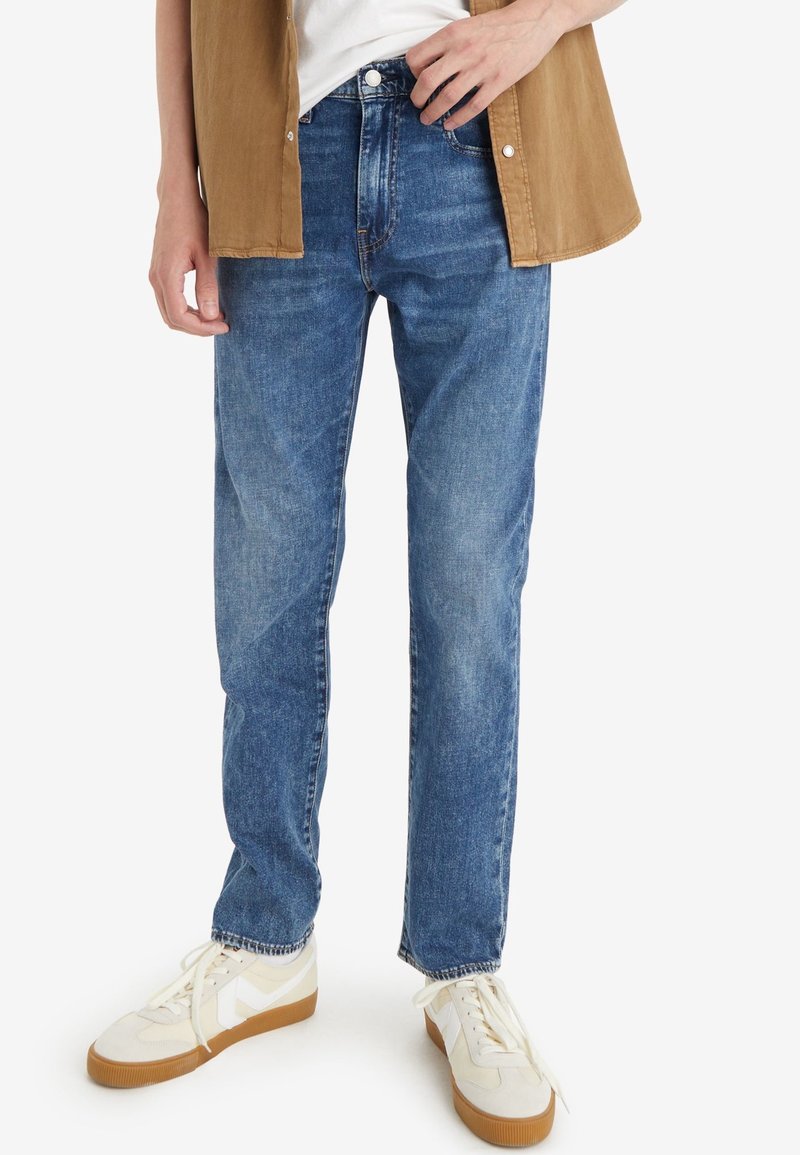 Personne portant un jean bleu droit, des baskets blanches avec semelles en gomme, une chemise blanche et une chemise marron ouverte à boutons.