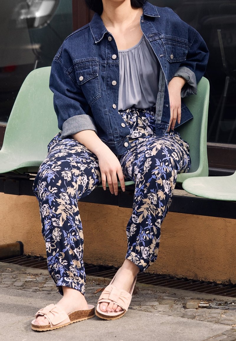 En denimjakke over en grå bluse, parret med bukser i blomsterprint i marineblå og lyserøde slidssandaler med en sløjfe detalje. Sidder på en grøn stol.