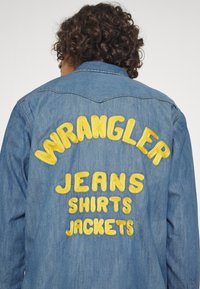 Persona con capelli ricci che indossa una giacca di denim blu con la scritta ricamata gialla "WRANGLER JEANS SHIRTS JACKETS" sulla schiena.