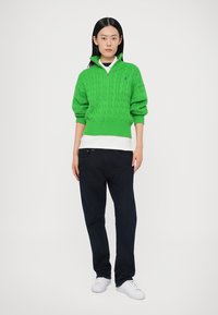 Green cable-knit sweater with a half-zip collar, worn over a white shirt, paired with dark denim jeans and white sneakers. Simple design.