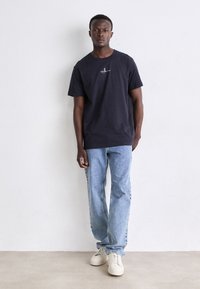 Navy cotton t-shirt with a round neckline and a small logo on the chest, paired with light blue straight-leg jeans and white sneakers.
