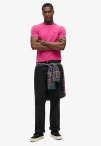 T-shirt di cotone rosa con maniche corte, pantaloni neri e una camicia di flanella a quadri legata in vita; abbinata a sneakers nere.