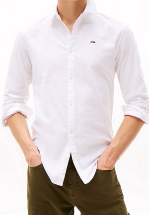 Mand iført en hvid langærmet skjorte med knappede manchetter, ærmerne rullet op, og olivengrønne bukser, hænderne i lommerne.