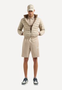 Giacca puffer beige con zip, polo color sabbia e pantaloni corti abbinati. Completata da un cappellino, con dettagli del logo, e scarpe nere.