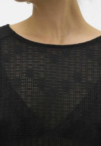 Närbild av en svart genomskinlig spetsblus med blommönster som bärs över huden, vilket visar linningen och övre bröstområdet.