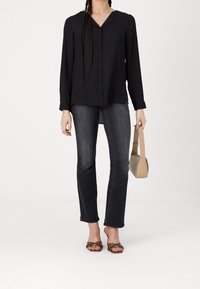 Blouse longue noire à manches longues avec un col en V, associée à un jean foncé. Sandales marron à enfiler avec des nœuds. Sac à main beige de forme rectangulaire.