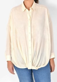 Cream-colored, lightweight button-up shirt with a collar, long sleeves, and a knotted front design. Made from a soft, sheer fabric.