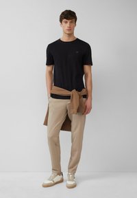 Schwarzes T-Shirt mit kurzen Ärmeln, kombiniert mit hellbeigen, taillierten Hosen. Ein brauner Sweatshirt ist um die Taille gebunden. Weiße und beige Turnschuhe runden das Outfit ab.