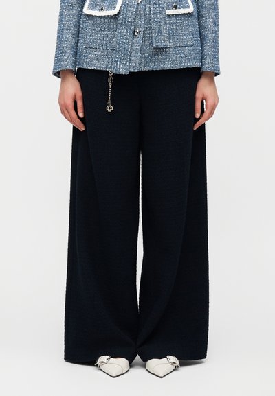 Μοντέλο με μαύρο παντελόνι φαρδιάς γραμμής, μπλε υφασμάτινο σακάκι με τσέπες στο μπροστινό μέρος και λευκά μυτερά παπούτσια με ασημένιες αγκράφες.