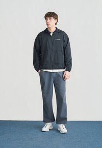 Junger Mann trägt eine schwarze Columbia-Jacke, blaue Hose und weiße Turnschuhe, steht mit den Händen in den Taschen auf einem blauen Teppich.