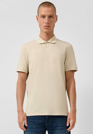 Homme aux cheveux courts portant un polo beige à manches courtes et un jean bleu foncé, debout devant un fond gris clair uni.