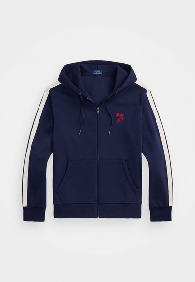 Sudadera con cremallera en color navy, con una gran capucha, bolsillos frontales y franjas blancas en contraste en las mangas. Presenta un logo rojo en el pecho.