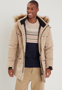 Veste matelassée beige avec une capuche en fourrure, dotée de poches avant et d'une fermeture éclair, portée sur un pull en tricot à motifs multi. 