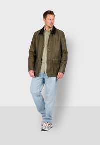 Brown leather jacket with a dark collar, front pockets, and button closure, worn over a light green shirt and light denim jeans.