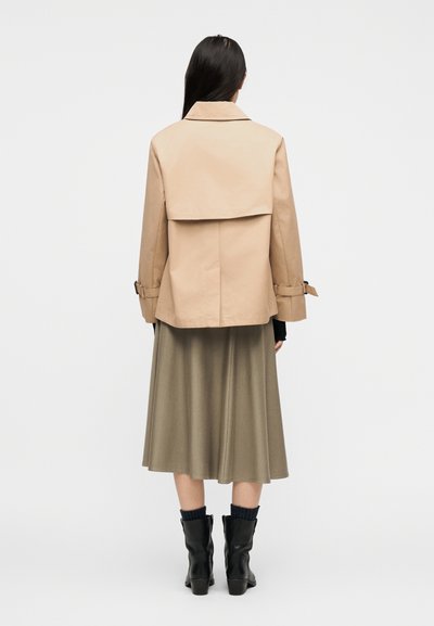 Γυναίκα που στέκεται με την πλάτη γυρισμένη, φορώντας μπεζ κοντό παλτό τύπου trench, ελιάς μέσης μέσης φούστα, μαύρες κάλτσες και μαύρα μπότες με τακούνι σε απλό φόντο.