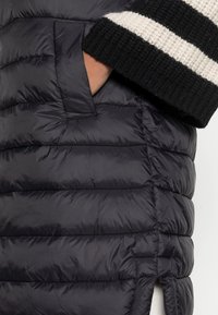 Czarna puchowa kurtka z poziomym pikowaniem, wyposażona w boczną kieszeń. Dzianinowe mankiety są czarne z białymi paskami. Miękka, gładka tekstura.