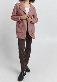 Personne portant un manteau rose texturé sur un haut blanc, un pantalon marron et des bottes noires épaisses, debout devant un fond clair uni.