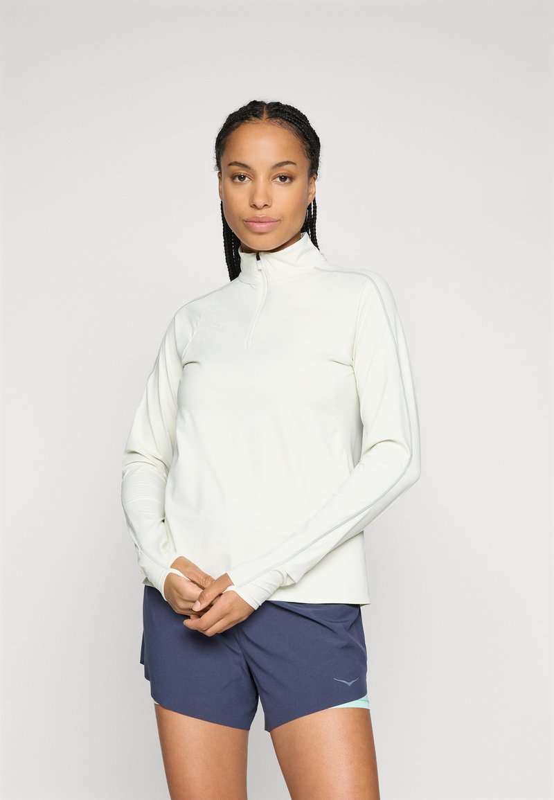 Woman with braided hair wearing a white long-sleeve zip-up top and navy blue athletic shorts, standing against a plain background.