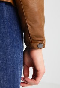 Main reposée à côté de jeans en denim bleu, portant un poignet de veste en cuir marron avec un bouton en métal portant l'inscription "ONLY" sur un fond uni.