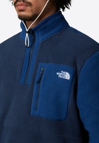Pullover in pile navale con zip parziale sul davanti, colletto alto, tasca sul lato sinistro del petto e logo ricamato, caratterizzato da una superficie morbida e strutturata.