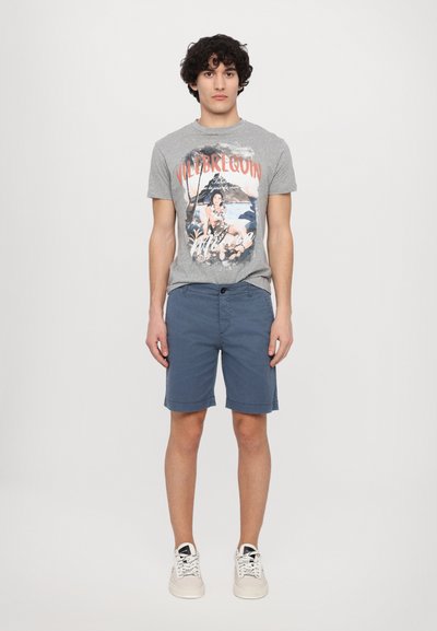 Camiseta gráfica gris con un estampado de playa, combinada con pantalones cortos azul marino. Modelo con zapatillas blancas, de pie frente a un fondo liso.