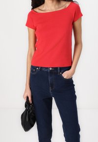 Červený bavlněný top s krátkým rukávem a off-shoulder designem, spojený s tmavě modrými džíny a držením černé kožené tašky.