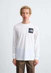 Langärmliges weißes T-Shirt mit einem schwarzen quadratischen Logo auf der linken Brust. Klassische Passform, Baumwollmaterial, weiche Textur, schlichtes Design.