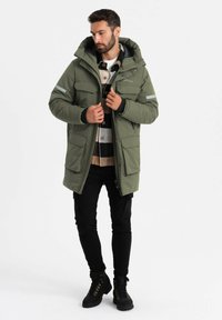 Giacca parka imbottita di colore verde oliva con cappuccio, grandi tasche frontali e dettagli riflettenti, indossata sopra una camicia a quadri, abbinata a pantaloni neri e stivali.