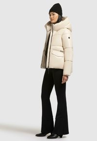 Giacca beige imbottita con colletto alto, zip frontale e due tasche laterali; indossata sopra pantaloni neri a zampa e un berretto nero.