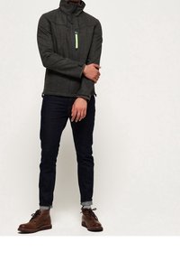 Veste zippée grise texturée avec col haute et détails de fermeture éclair vert fluo. Le modèle porte un jean foncé et des bottes à lacets marron.