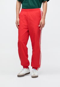 Rode sportieve broek met witte zijstrepen, elastische tailleband en ritsen bij de zakken. Gemaakt van een lichtgewicht stof voor comfort.