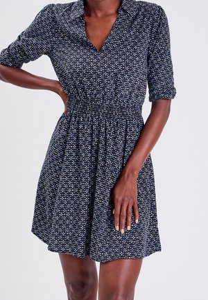 Kobieta ubrana w granatową sukienkę z białym geometrycznym kwiatowym wzorem, z elastyczną talią i podwiniętymi rękawami, stojąca na tle jednolitego tła.