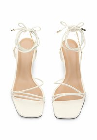 Valkoiset korkokengät, joissa on neliömäinen kärki, ohuet hihnat, solmittava nilkkaketju ja kultaketjuakseli. Tasainen mattapinta.