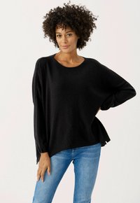 Schwarzer Strickpullover mit lockerer Passform, runden Halsausschnitt und überschnittenen Schultern. Kombiniert mit blauen Denim-Jeans, die eine glatte Textur und seitliche Schlitze aufweisen.