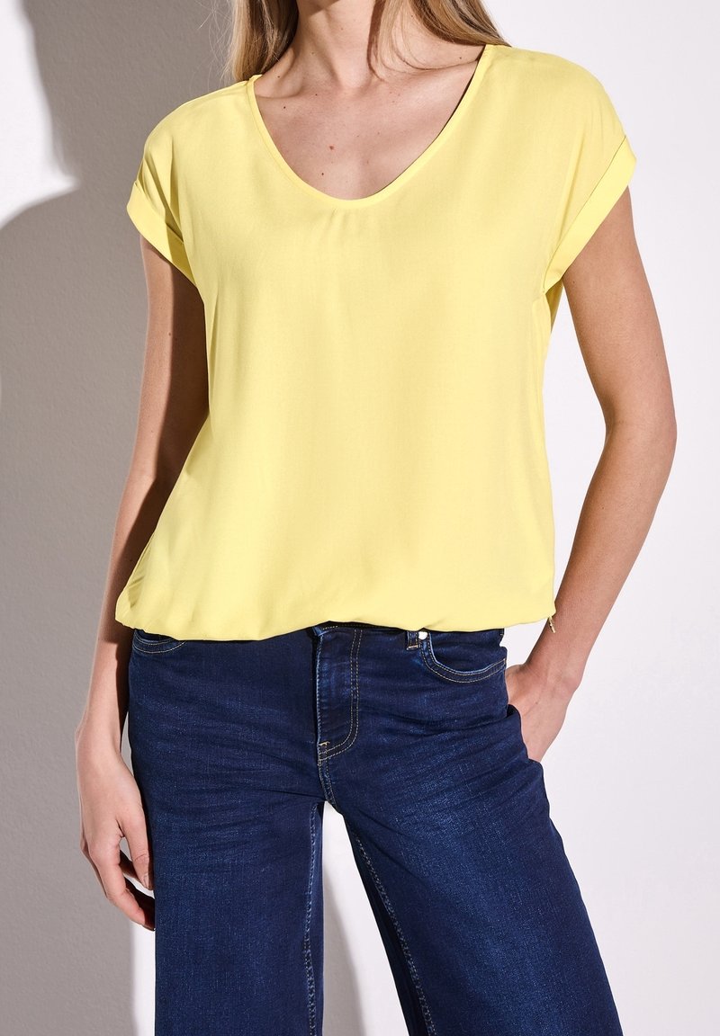 Blouse jaune à manches courtes avec un décolleté en V profond, fabriquée dans un tissu lisse, associée à un jean en denim bleu foncé avec un design classique à cinq poches.