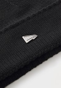 Crna pletena kapa s teksturiranim finišem, koja ima mali srebrni metalni logo na prednjoj strani.
