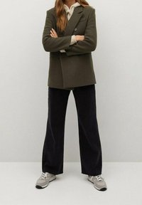 Olivově zelený dvouřadý blazer přes krémový svetr, sladěný s tmavými širokými kalhotami a šedými teniskami. Neutrální barvy, strukturovaný design.