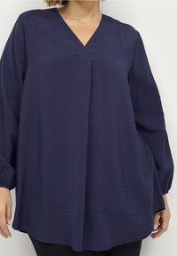 Blouse texturée bleu marine avec un décolleté en V, des manches longues et une coupe décontractée. Présente des plis subtils et un ourlet arrondi.