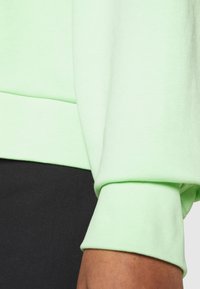 Tejido de sudadera verde claro con puños acanalados, textura suave y corte relajado. Detalles de costura visibles en las mangas y el dobladillo.