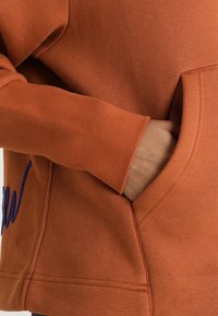 Orange sweatshirt med en strukturerad tyg, med en stor ficka och en blå broderad detalj. Manschetten är utrustad med en slät söm.